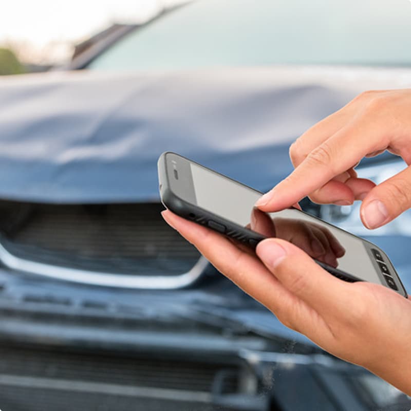 Déclaration d'accident auto sur smartphone après collision Personne utilisant un smartphone devant une voiture au capot froissé après un accident de la route.