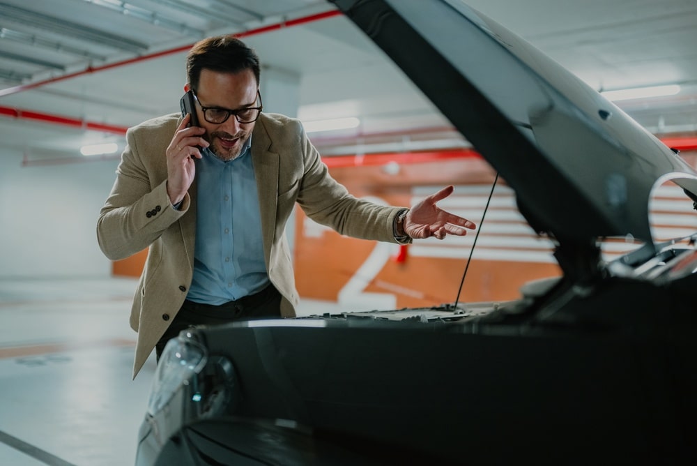Panne voiture parking : homme appelle l'assistance routière Homme frustré au téléphone devant le capot ouvert de sa voiture en panne dans un parking souterrain.