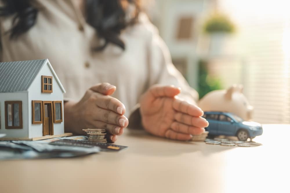 Mains protégeant des miniatures de maison, voiture et argent pour symboliser la gestion de patrimoine et l'assurance.