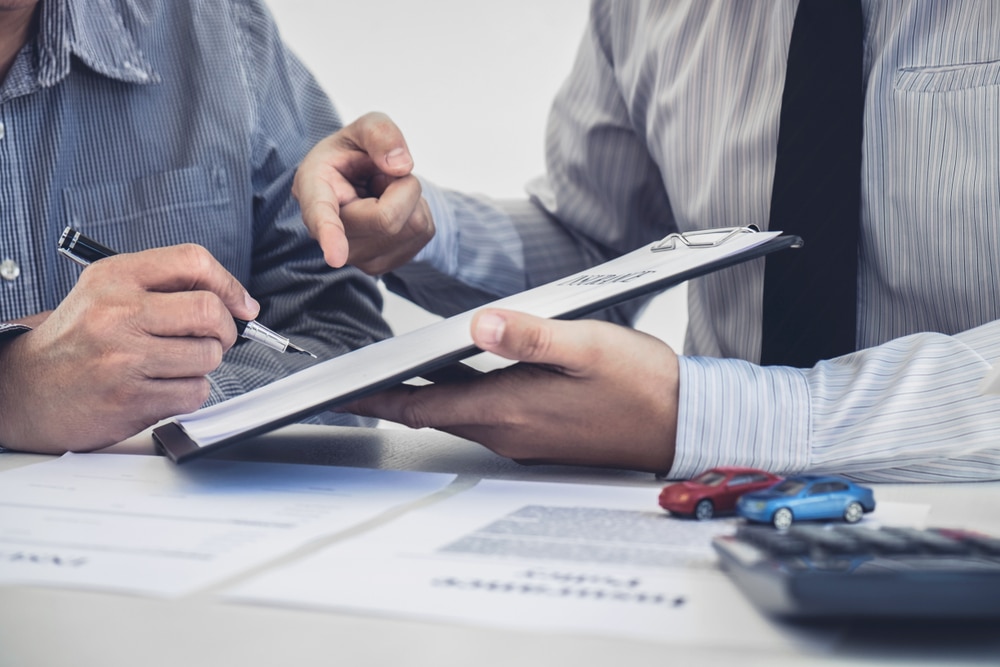 Assurance auto : signature d'un contrat avec un conseiller Un homme signe un contrat d'assurance auto présenté par un conseiller, petites voitures et calculatrice sur le bureau.