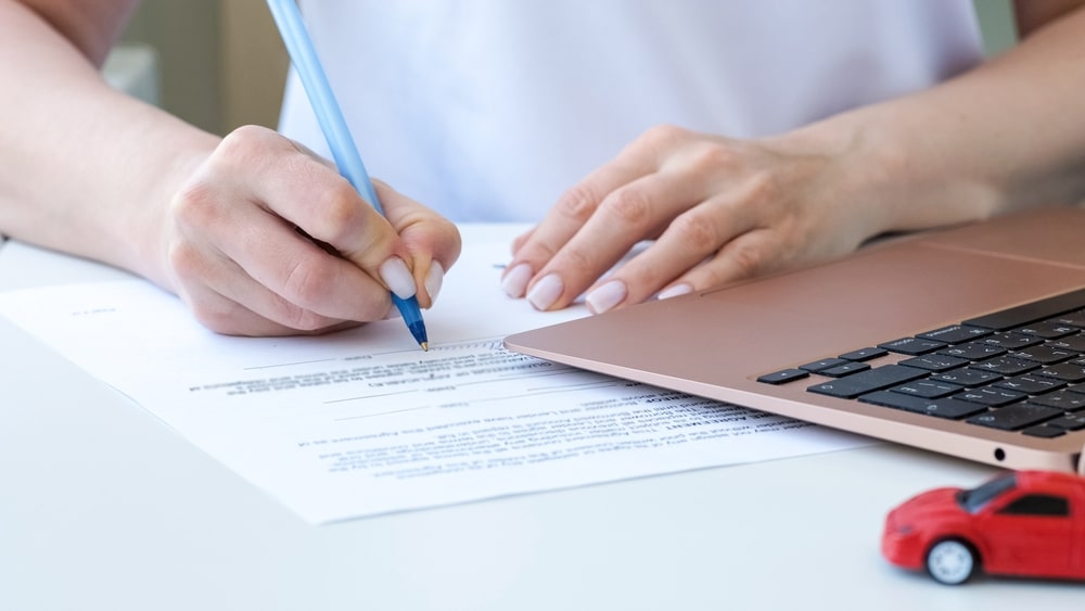 Main signant un contrat d'assurance auto à côté d'une voiture rouge miniature et d'un ordinateur portable.