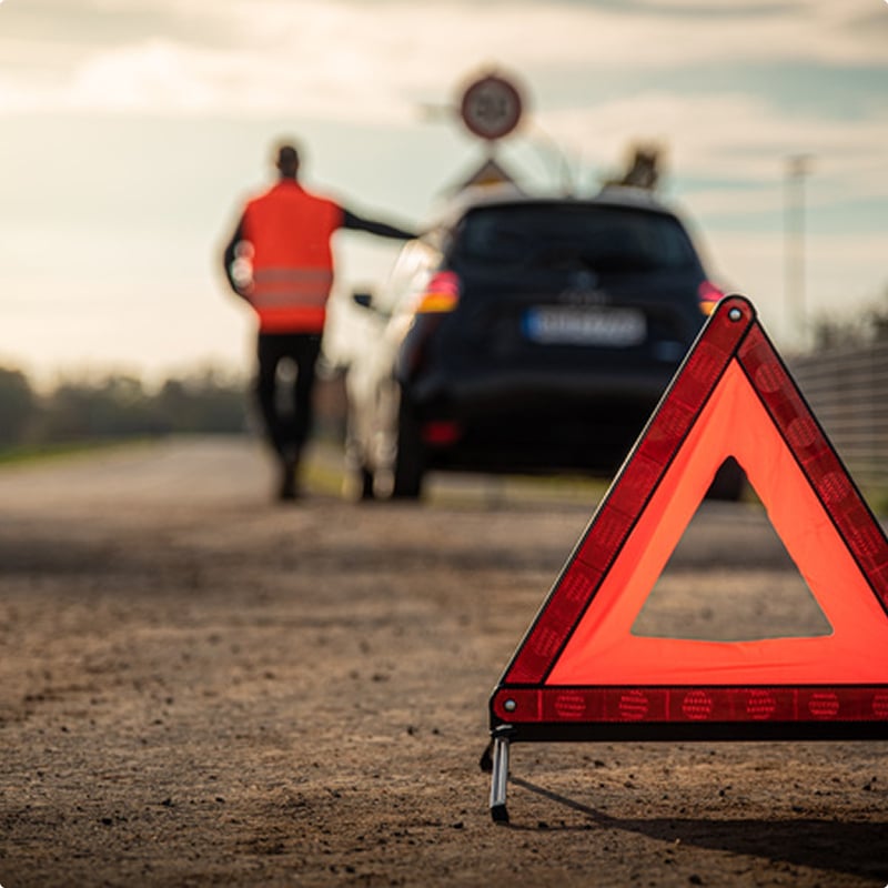 Panne de voiture : utiliser le triangle de signalisation Triangle de sécurité au premier plan, voiture en panne et conducteur en gilet orange sur le bas-côté d'une route.